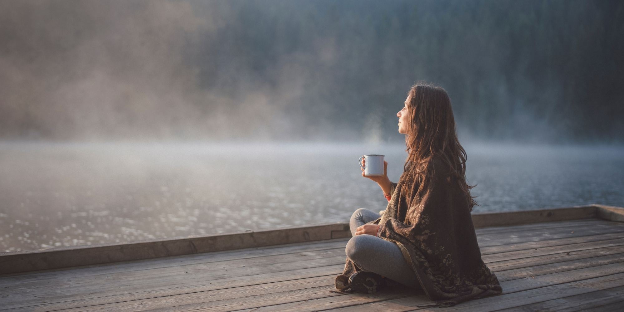 A woman sitting cross legged on a deck by a mimsty lake, with a steaming cup in her hand.