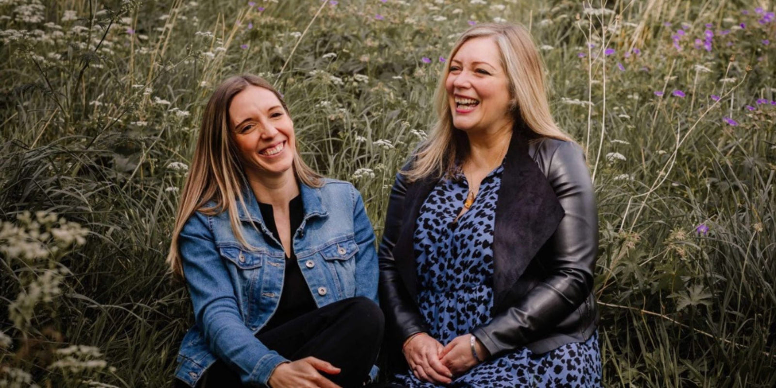 Two women smiling and laughing while sitting outdoors in a field of wildflowers. One woman wears a blue denim jacket over a black top, and the other wears a black leather jacket over a blue patterned dress.