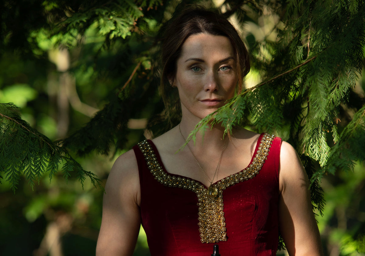 A woman wearing a sleeveless burgundy top with ornate gold embroidery around the neckline, standing outdoors among green trees.