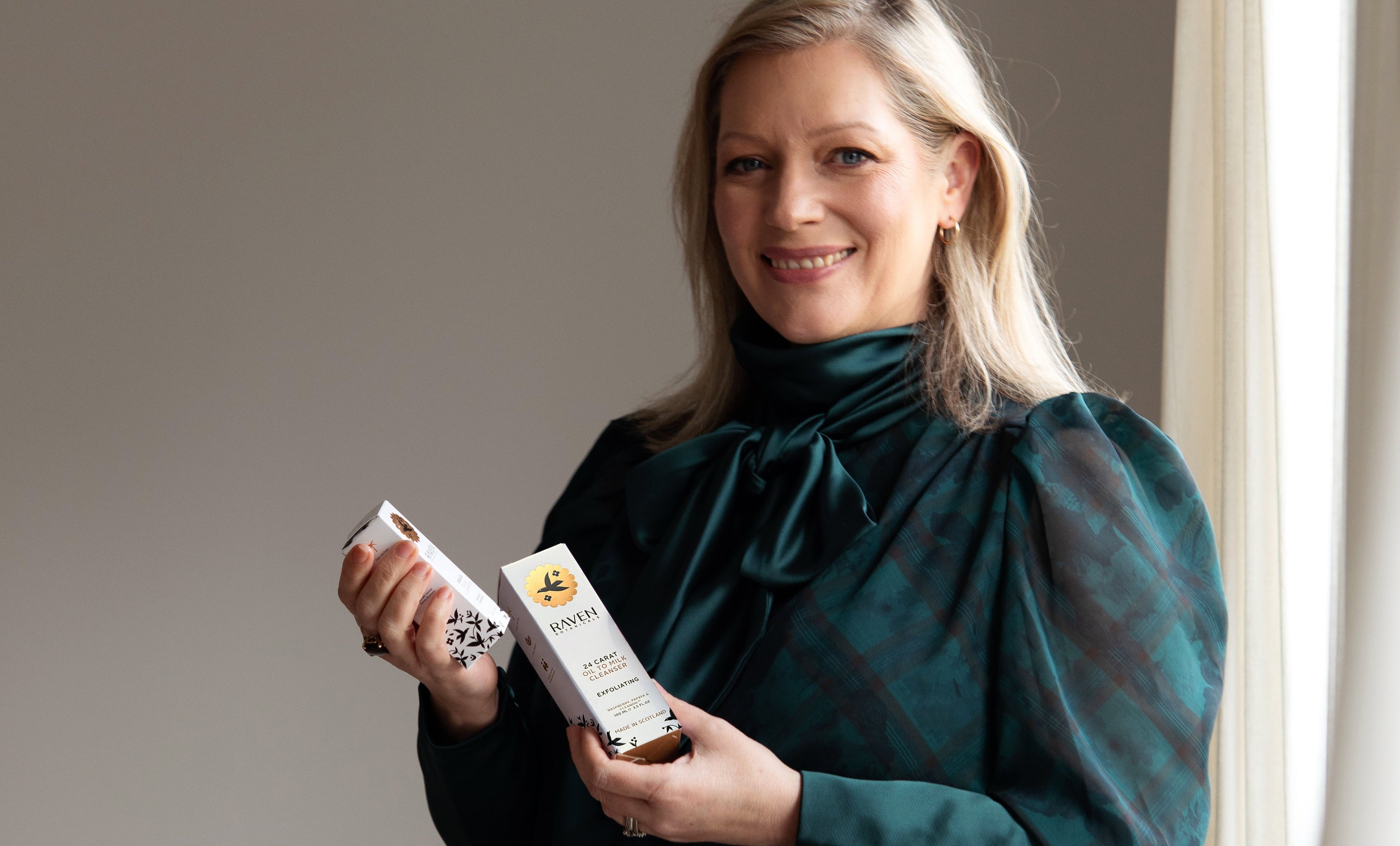 Woman holding two boxes containing Raven Botanicals products against a neutral background
