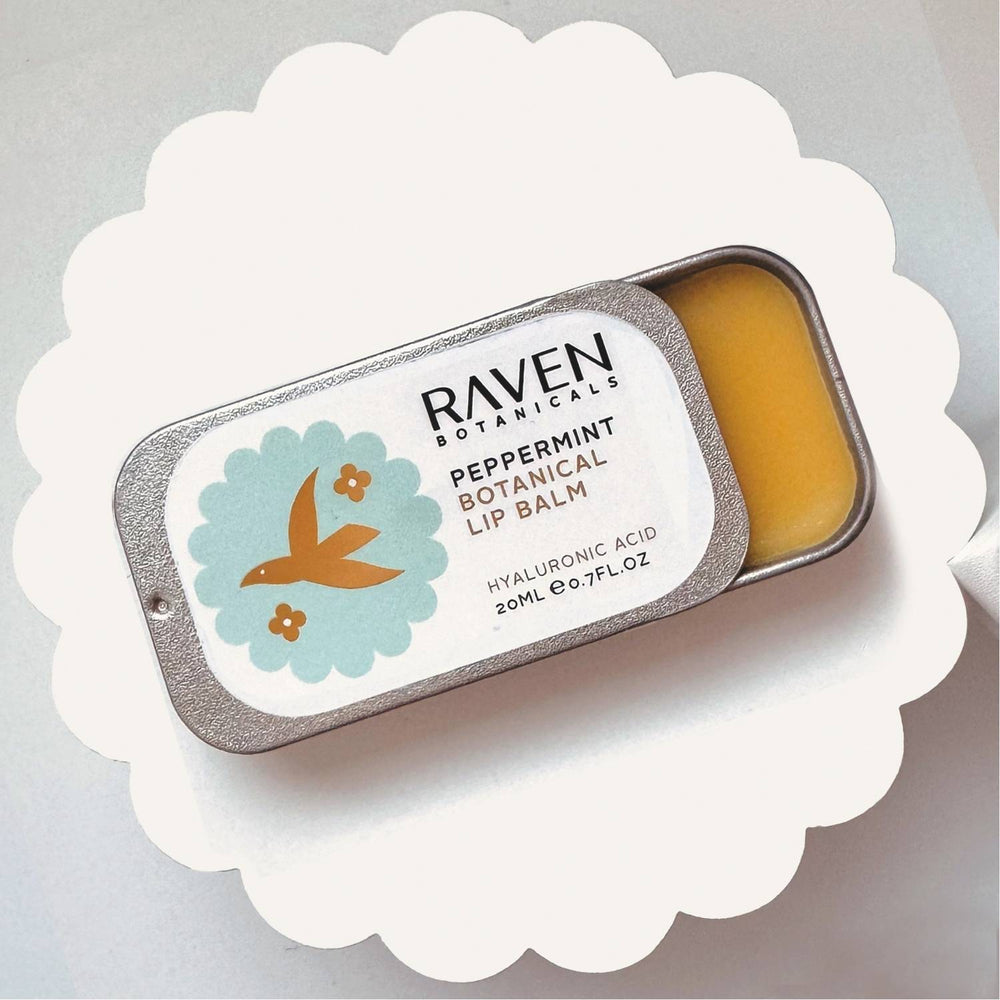 A small open container of Raven Botanicals Botanical Lip Balm in Peppermint flavour, with a label showing a bird and peppermint flowers.
