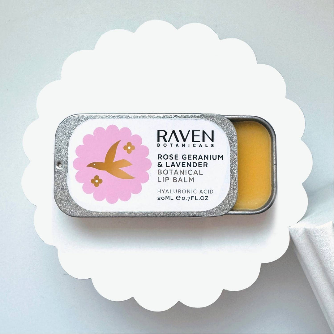 A small open jar of Raven Botanicals Rose Geranium Botanical Lip Balm on a white dome, with a label that includes the product name and a pink flower illustration.